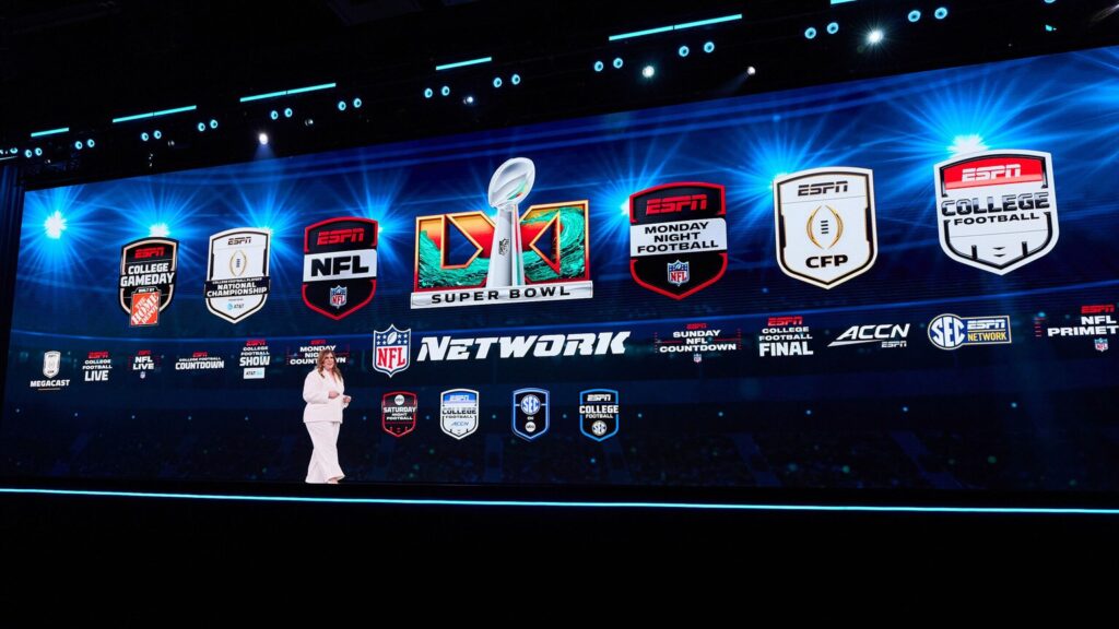 Stage with a large screen showing ESPN NFL Network logos and Super Bowl graphic; a presenter in white stands on a dark stage.