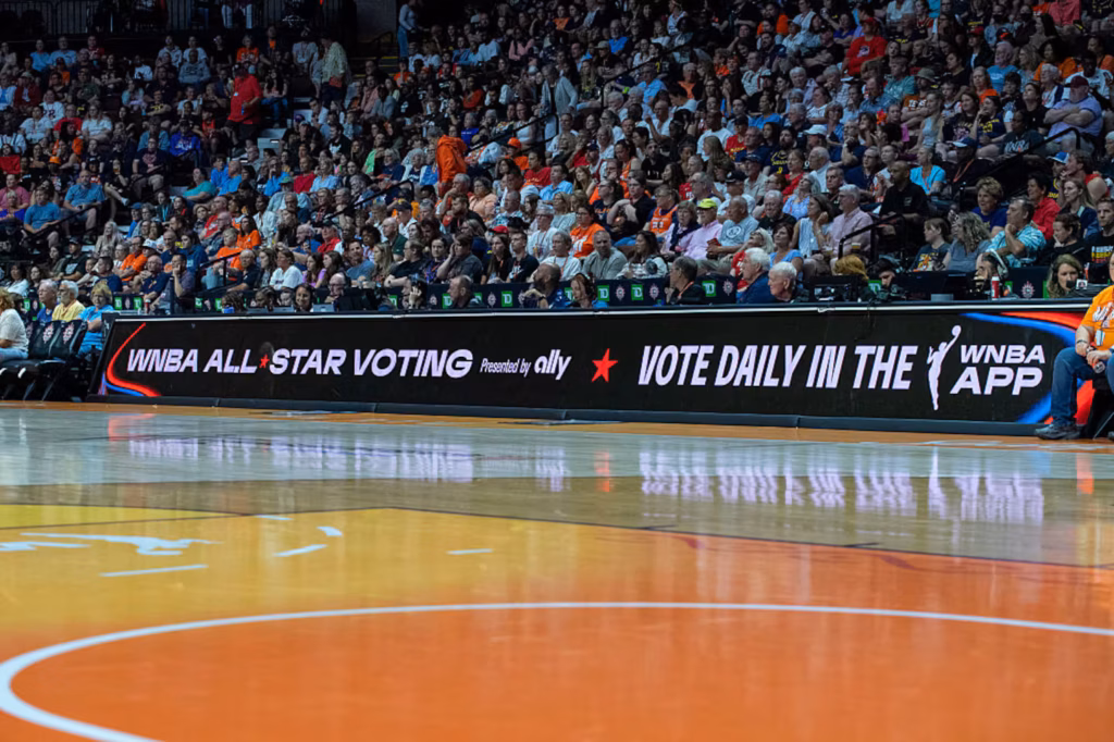 Crowd in a basketball arena behind a banner that reads 'WNBA All-Star Voting' and 'Vote daily in the WNBA app'.