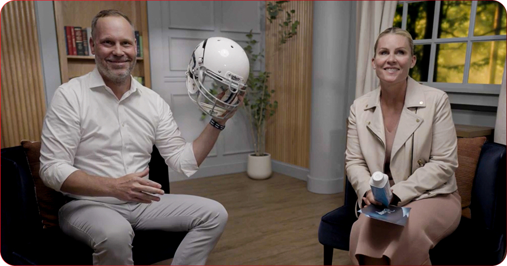 Two people sit in a cozy studio living room: man in white shirt holds a white football helmet, woman in beige jacket holds a brochure and a spray bottle, smiling at the camera.